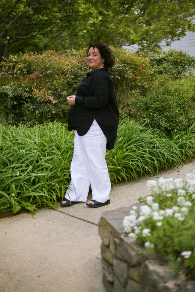 Alison of Wardrobe Oxygen standing in profile but is looking at the camera. She is wearing the Universal Standard Dawn tunic, a high-low hem button down linen shirt tunic, over a pair of white linen wide leg pants. On her feet are Birkenstock sandals.