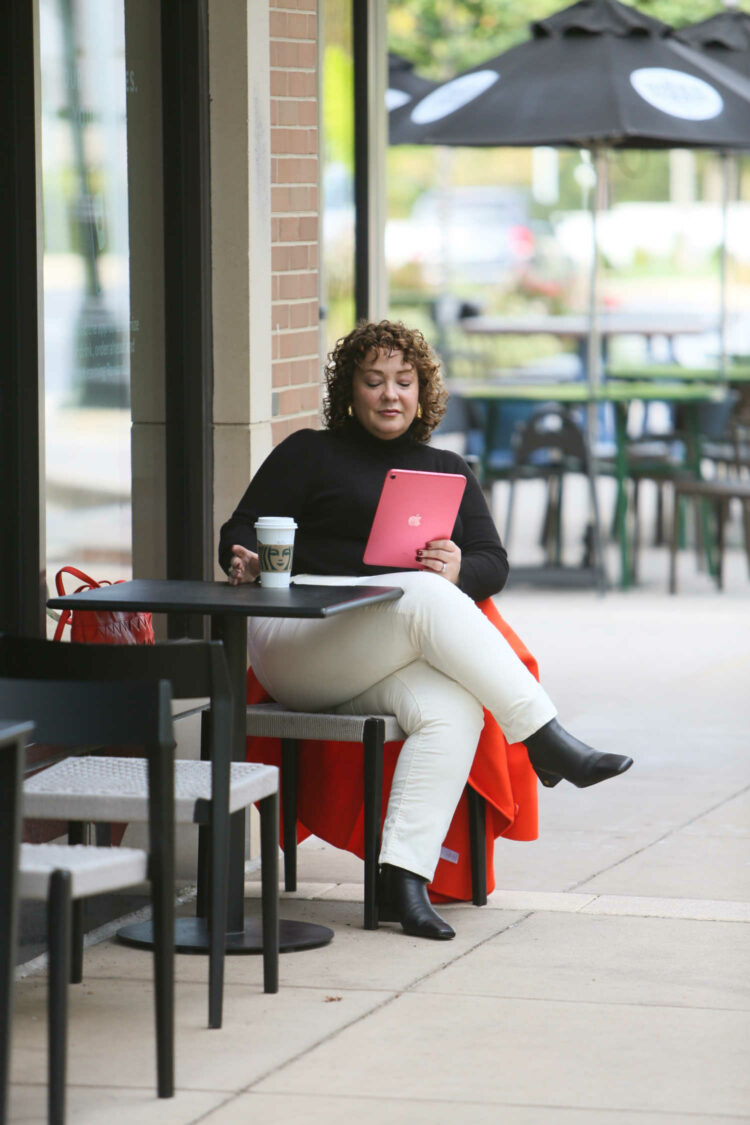 Alison Gary of Wardrobe Oxygen in a Talbots black turtleneck sweater and ivory corduroy jeans sitting outside a coffee shop reading from an iPad while drinking a latte