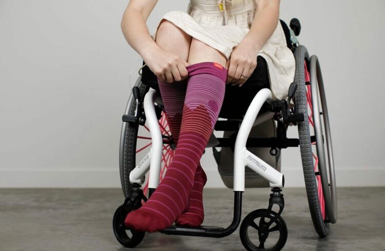 a woman in a wheelchair pulling up striped over-the-calf compression socks from Vim & Vigr