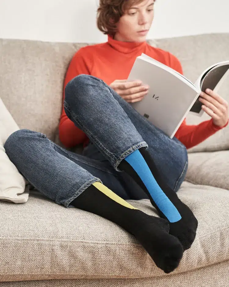 A woman sitting on a couch reading a book wearing black, yellow, and blue patterned compression socks from Ostrichpillow