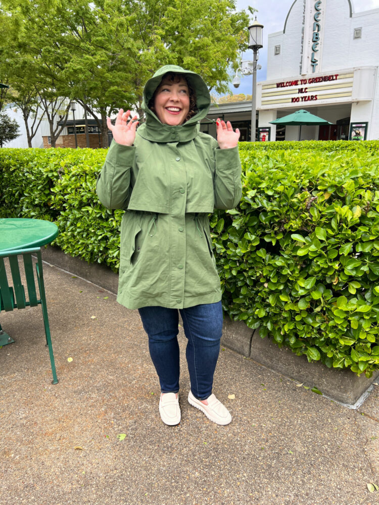 Woman standing in Roosevelt Center in historic Greenbelt, Maryland. She is in from of the Greenbelt Cinema, wearing the Universal Standard Precision Parka in green with dark slim jeans and ivory Vionic loafers.