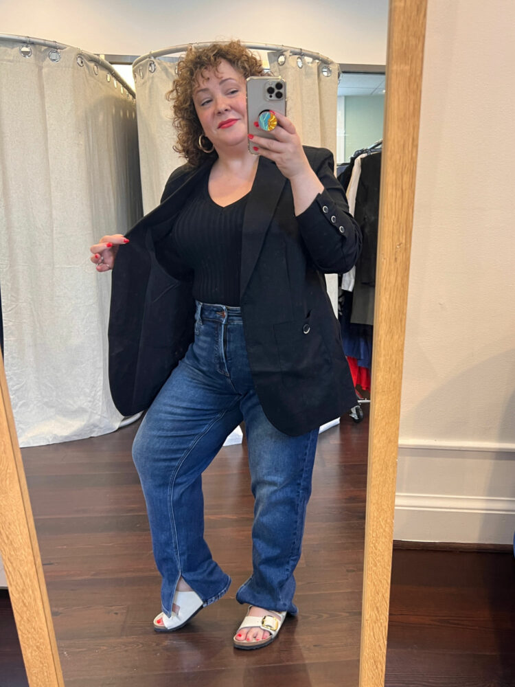 Alison Gary of Wardrobe Oxygen at a Universal Standard trunk show. She is taking a mirror selfie as she tries on a black linen blazer, black v-neck sweater vest, and jeans