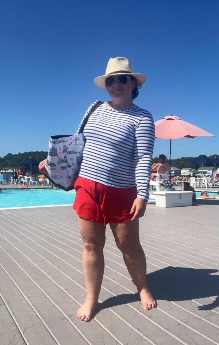 Alison Gary in a striped Lands' End rashguard with red Universal Standard Sunny active shorts, a straw hat, and Scout bags DC Deano tote. She is at the pool