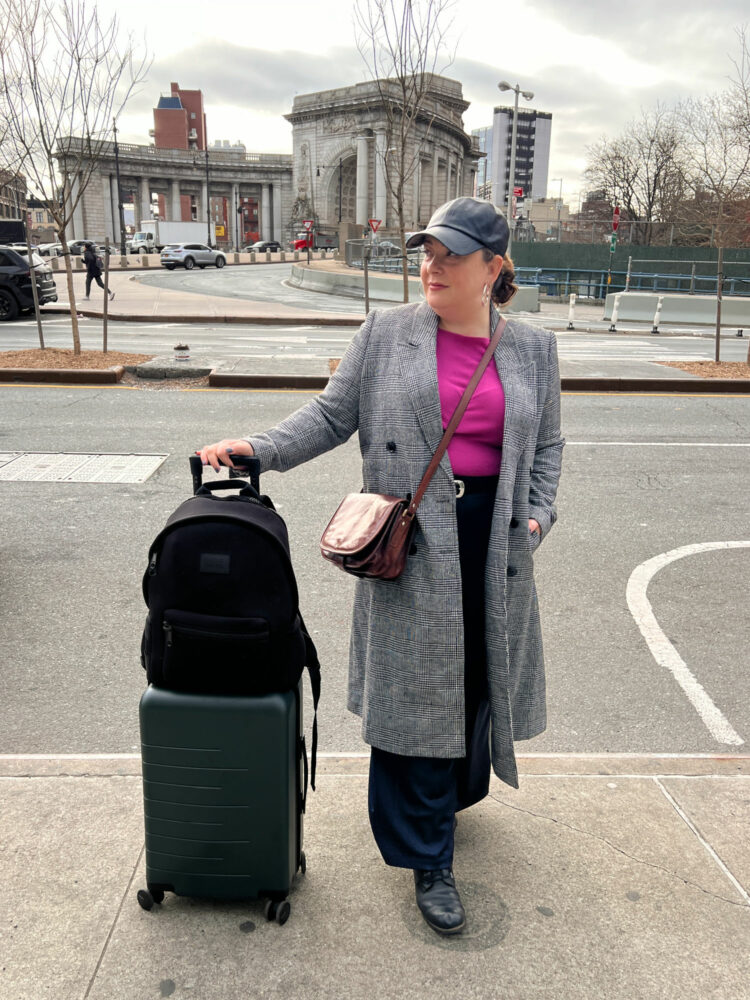Alison Gary of Wardrobe Oxygen outside an NYC hotel holding onto a Quince rolling suitcase that has a Dagne Dover Dakota backpack on top of it.