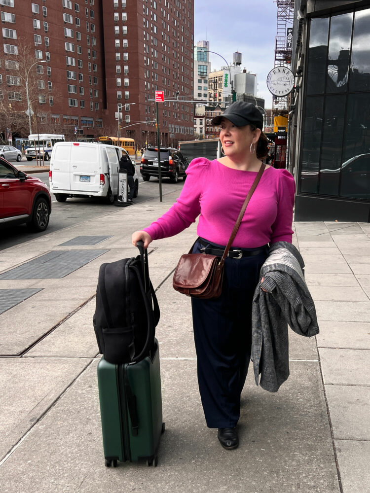 Alison Gary of Wardrobe Oxygen outside an NYC hotel holding onto a Quince rolling suitcase that has a Dagne Dover Dakota backpack on top of it.