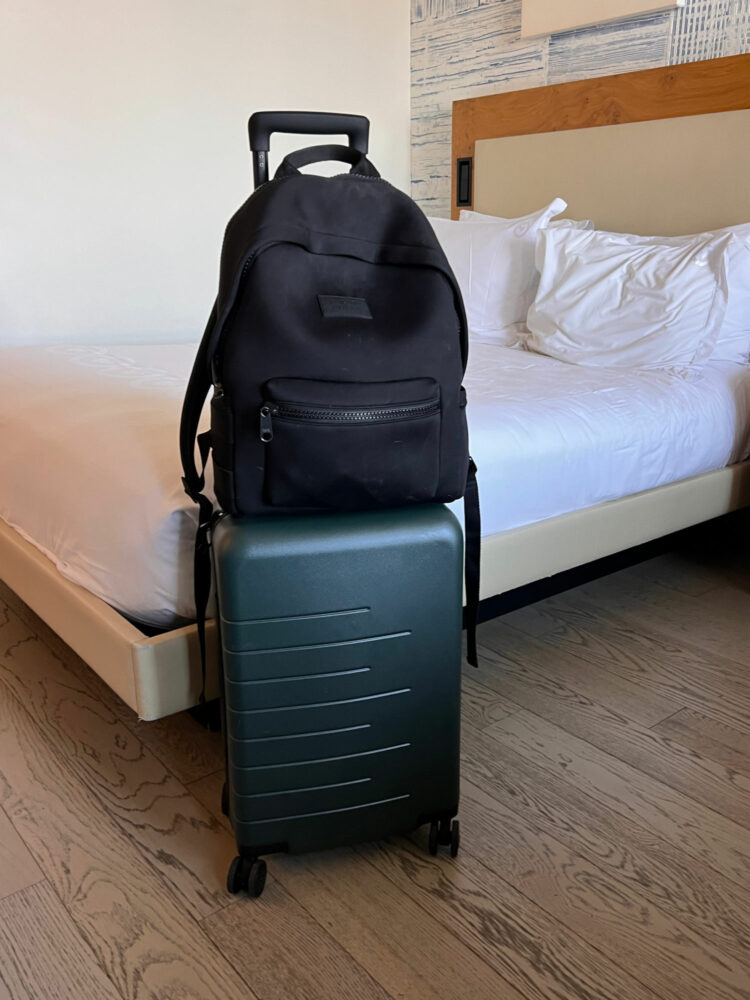 The Quince 20" Expandable Carry-on Spinner suitcase with the Dagne Dover Dakota backpack on top in a hotel room