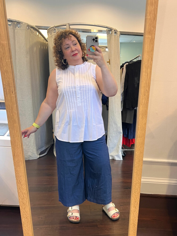 Alison wearing a white sleeveless cotton popover with pintucks and covered buttons worn untucked over chambray wide-leg ankle pants