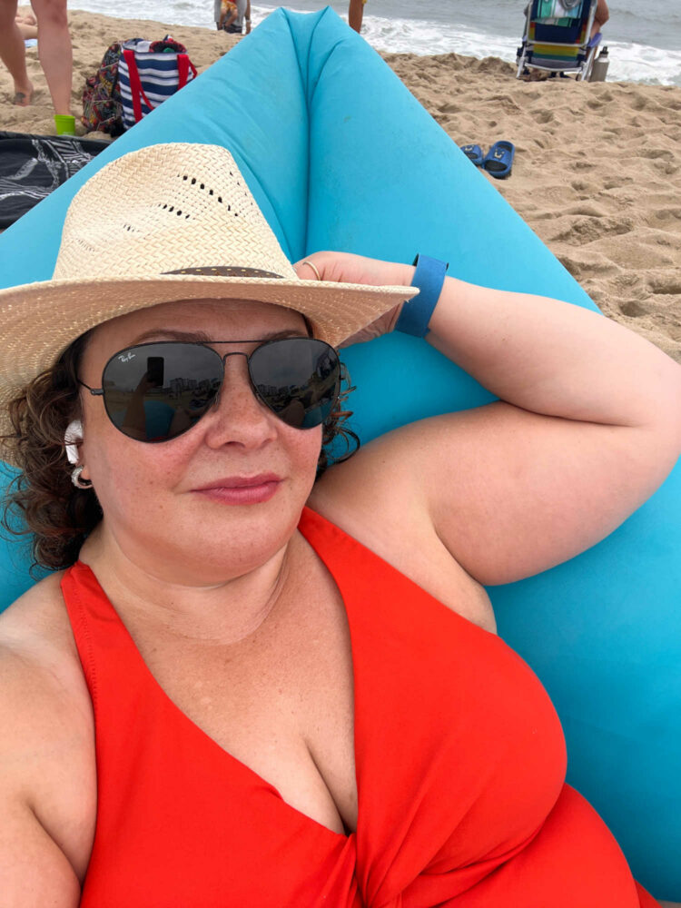 Alison at the beach lying in a fatboy lounger, wearing a straw hat and black aviator sunglasses and the Universal Standard The Swimsuit in Sun Fire.