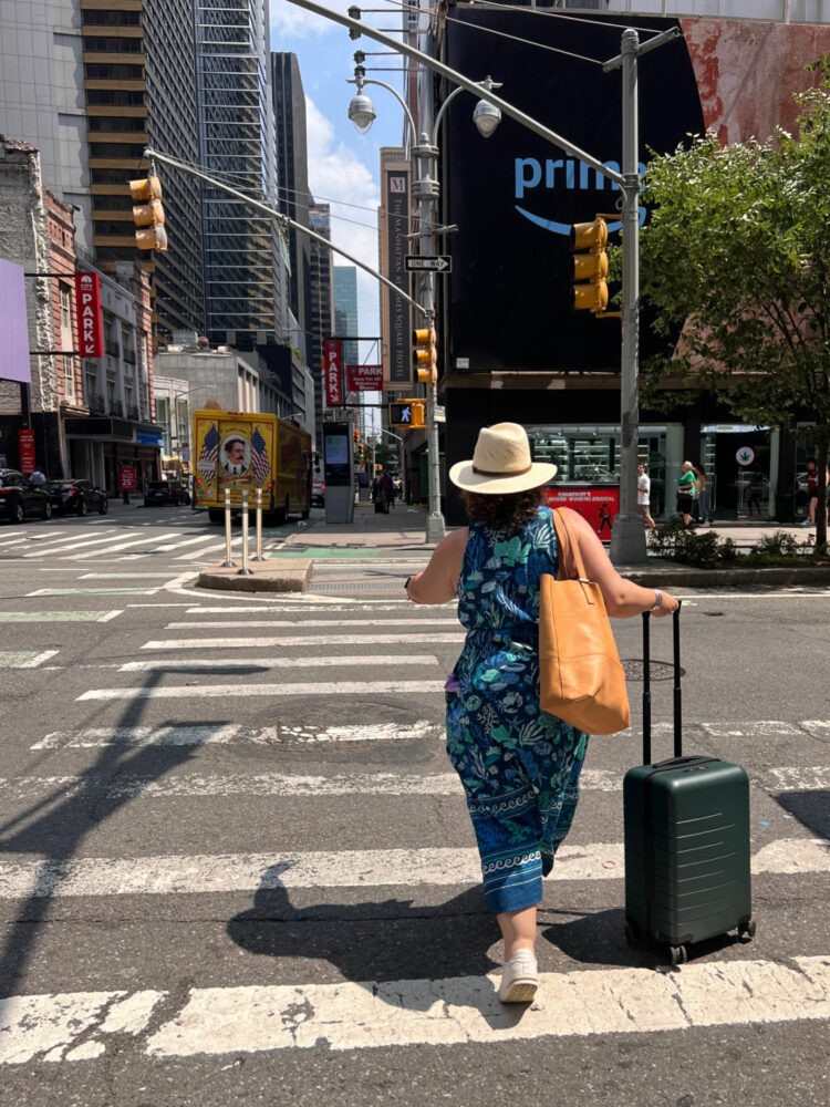 Alison of Wardrobe Oxygen crossing a street in NYC rolling a suitcase and wearing a blue printed shirtdress from Printfresh