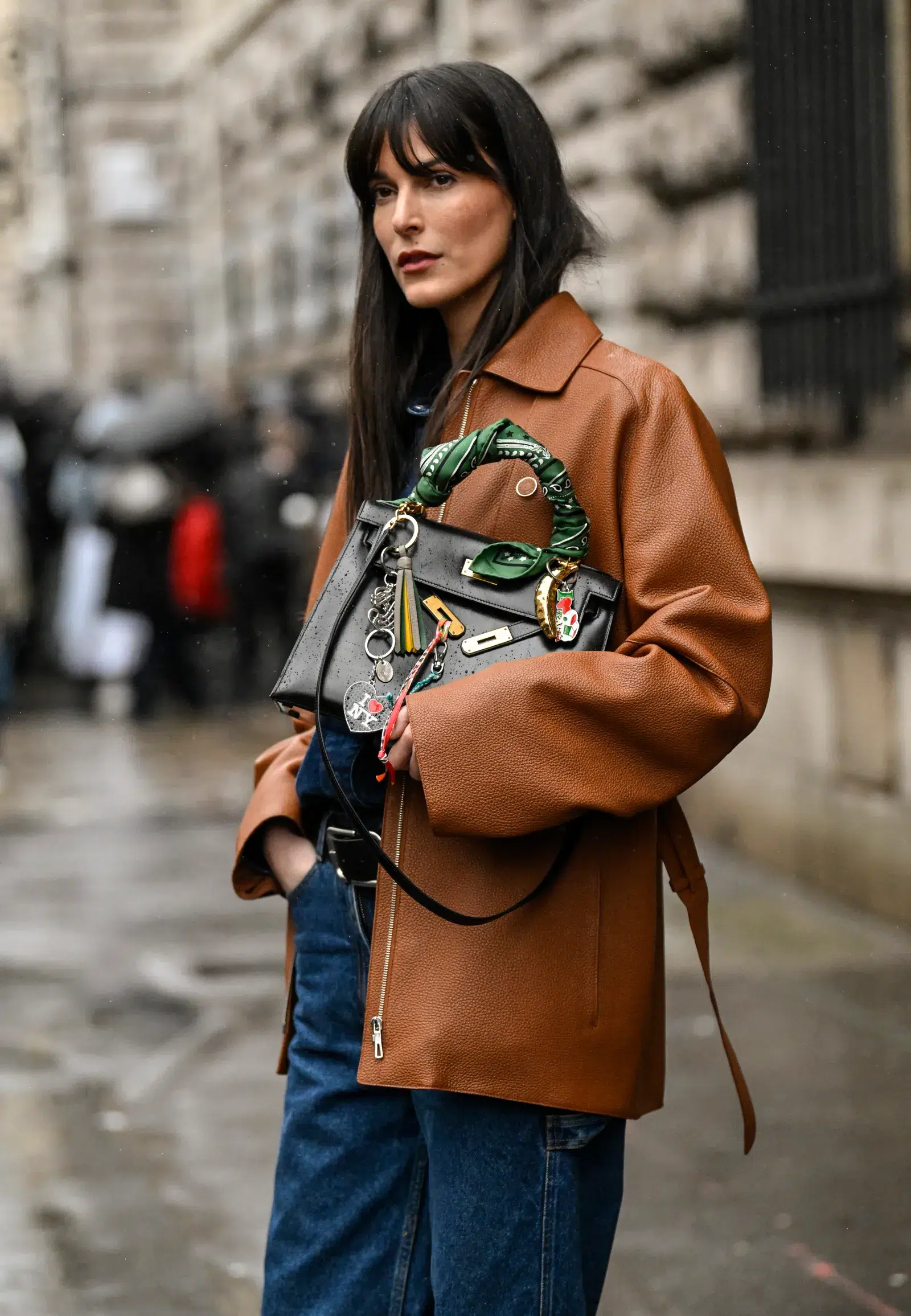 woman in a brown leather coat holding a leather handbag embellished with chains, a scarf, and bag charms