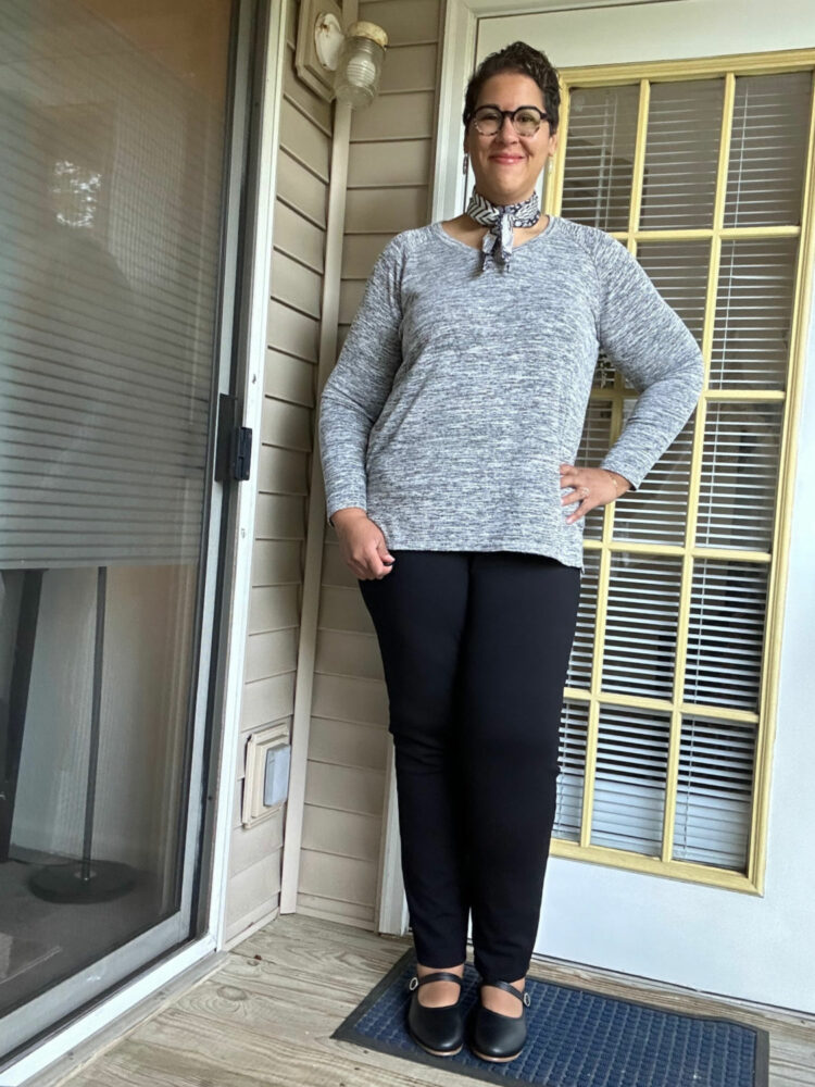 woman in a gray marled tunic sweater and black pants styled with black leather Mary Jane flats and a scarf at the neck