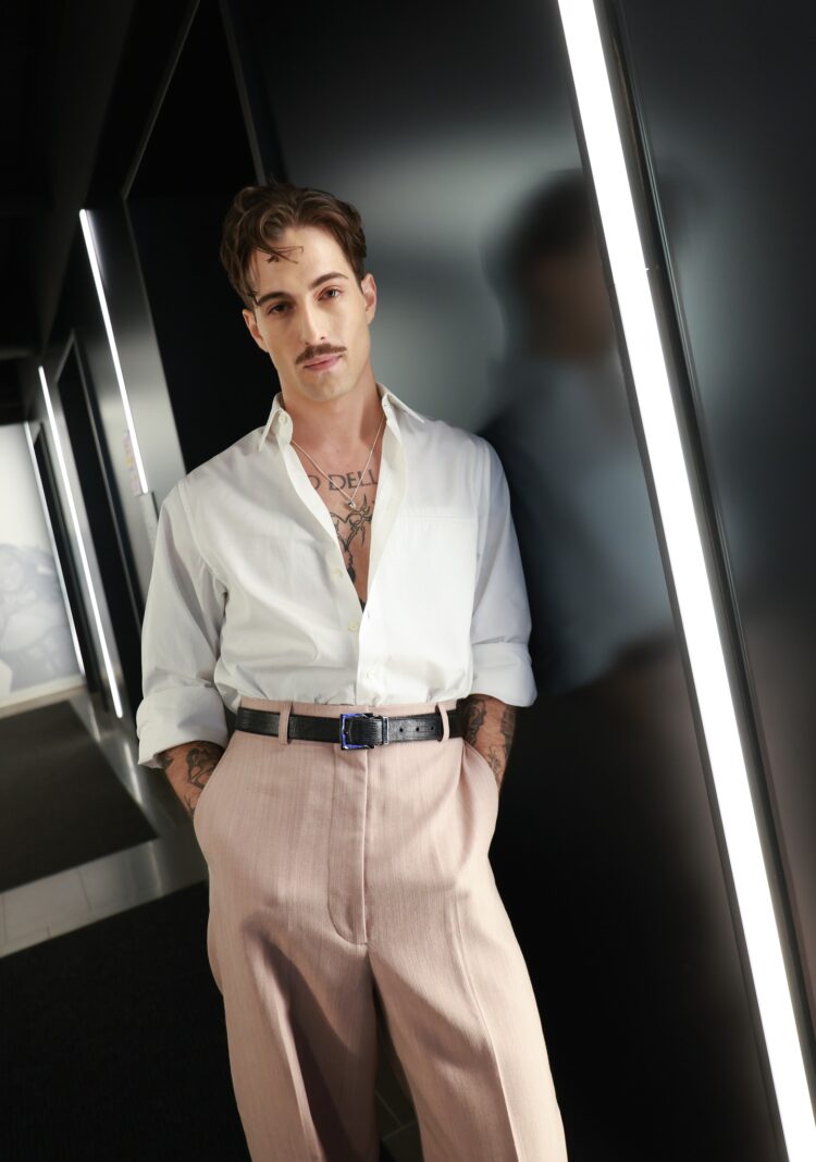 Damiano David in a white button-front shirt unbuttoned low and sleeves rolled to show his tattoos. Shirt tucked into tan trousers with a black belt; he has his hands in his pockets and is leaning against a wall staring at the camera.