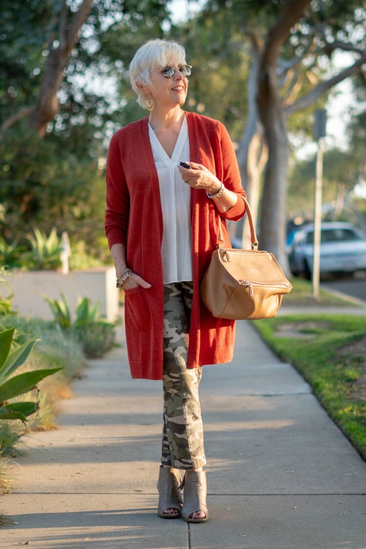 Susan from the blog Une Femme in a dark orange long cardigan, white v-neck shell, camo pants, and a tan handbag.