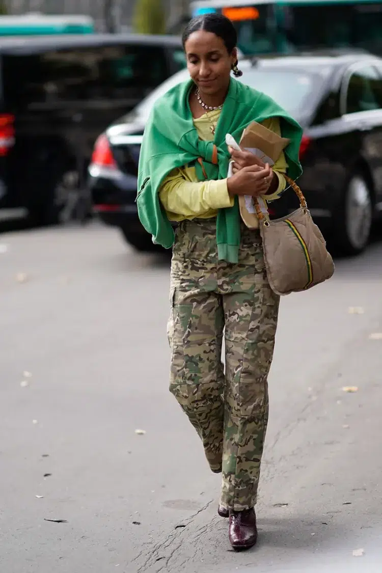 woman wearing camo pants with a pale celadon sweater and a kelly green sweater tied over her shoulders, holding a classic handbag