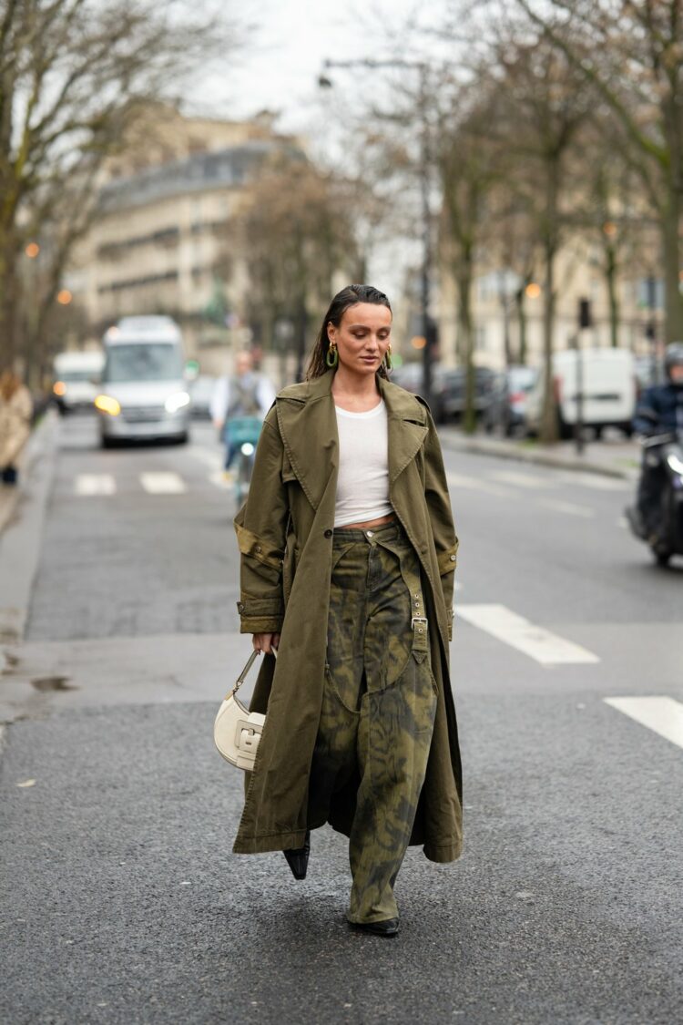 Photo by KaraStreetStyle from Paris Fashion Week Autumn/Winter 2024 of a woman in an olive trench, fitted white tee, and high waisted full leg camo pants