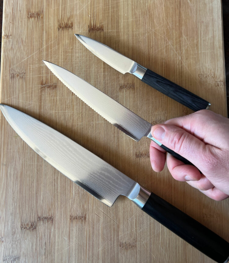 three kitchen knives on a wooden cutting board. One is being held in the hand of a man