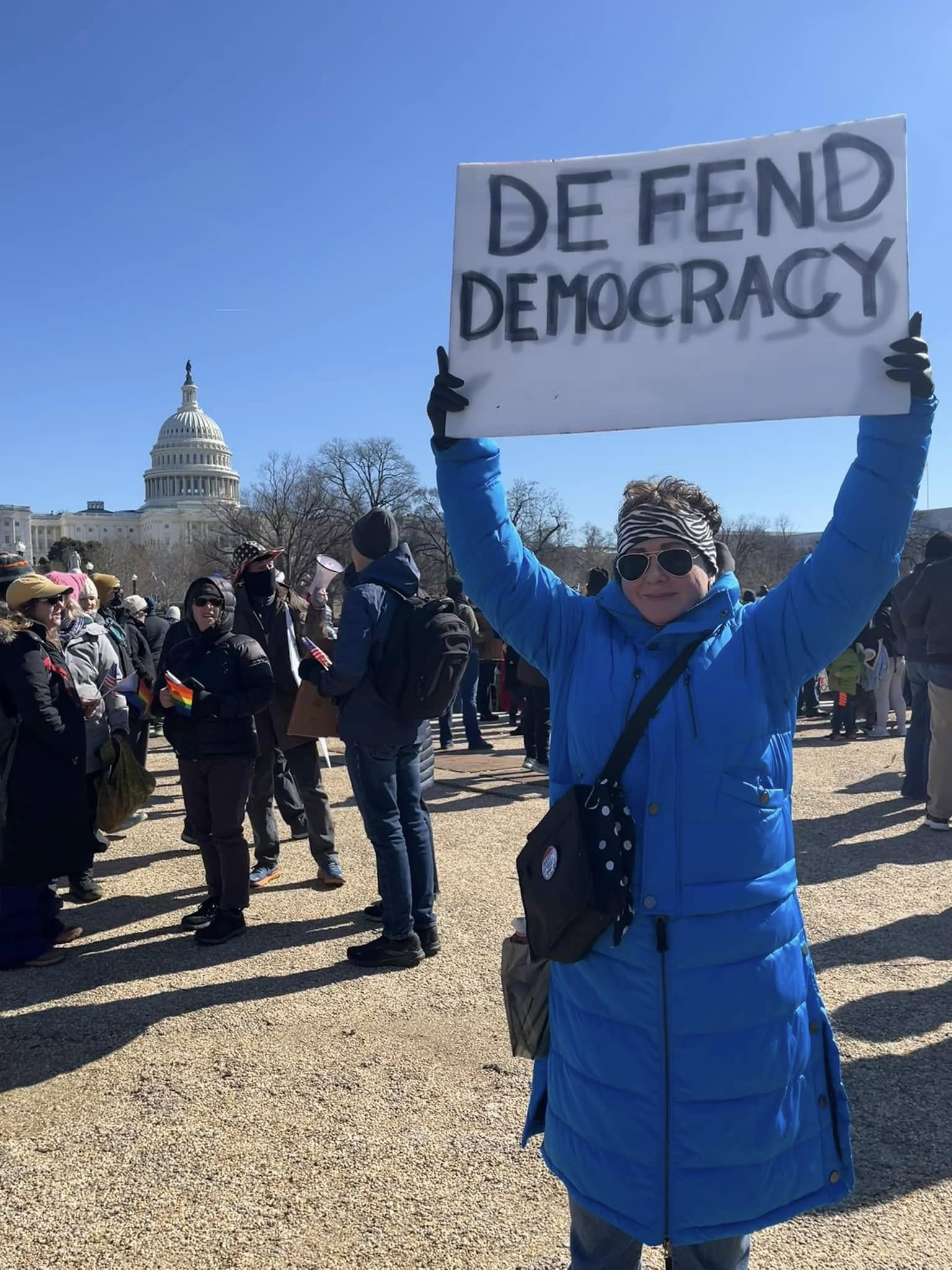 Alison at the President's Day Protest in 2025 wearing a long blue winter coat and holding a sign that says Defend Democracy