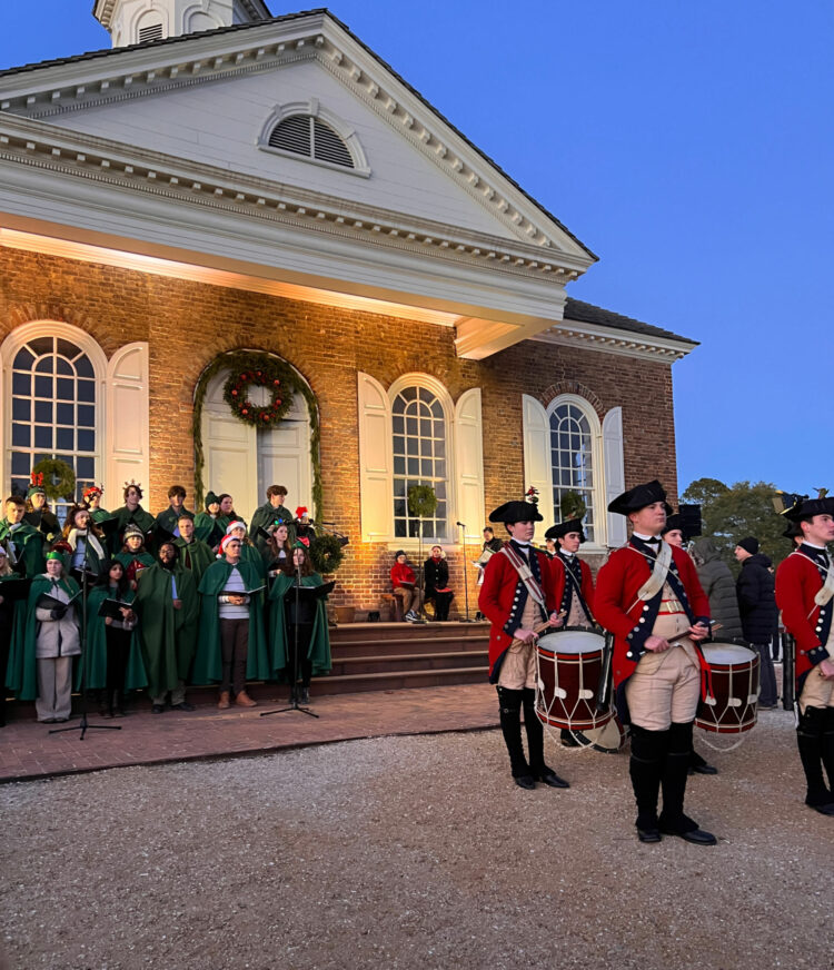 The Colonial Williamsburg Courthouse with a choir on the steps and a fife and drum corps performing in front. It is sunset and the Courthouse has a wreath on the door as this was a visit to Williamsburg in December